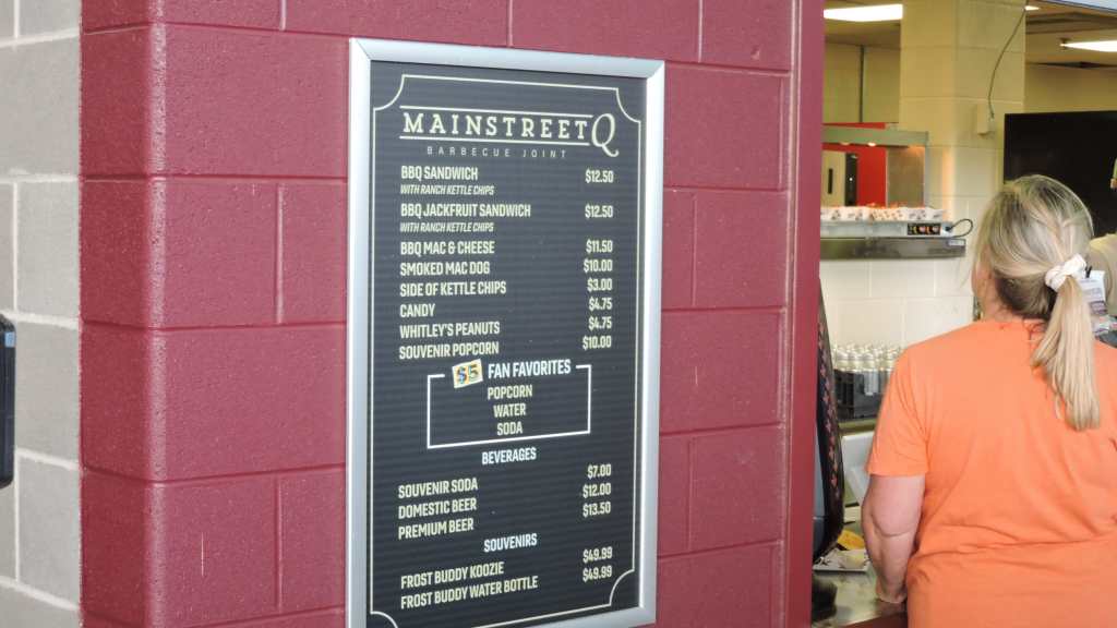 Menu board at a barbecue joint featuring various food and beverage options with prices, and a customer standing in front of it.