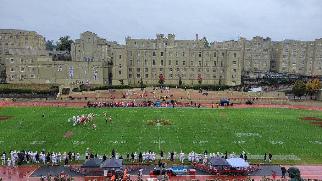 A view of a college football game in progress, featuring players on the field with a historic building in the background and spectators along the sidelines.