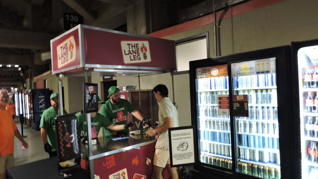 A concession stand named 'The Lane Leg' at a sports venue, featuring two staff members behind the counter, with a customer making a purchase. A beverage fridge is visible in the background, filled with various drinks.