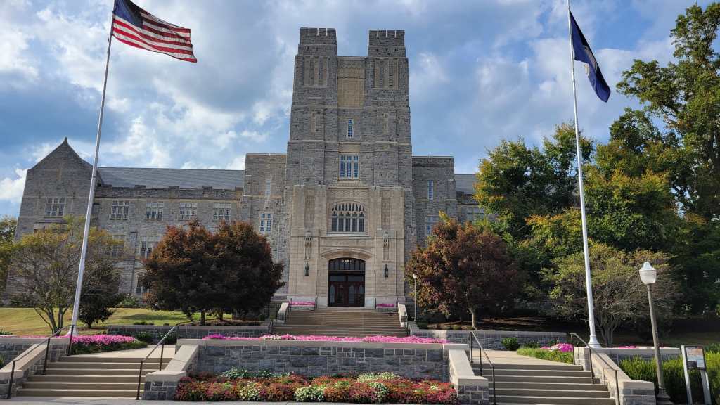 Exterior view of a large stone academic building with a tall tower, flanked by U.S. and state flags, surrounded by landscaped grounds with colorful flowers.