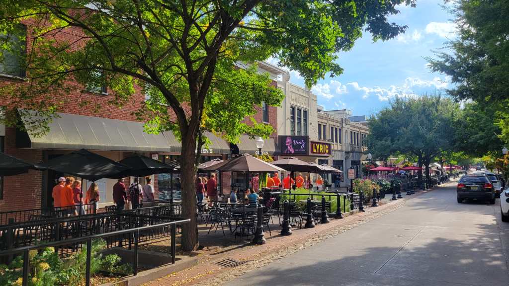 A lively street scene featuring cafes with outdoor seating, shaded by trees, and people walking along the sidewalk. Brightly colored signage and buildings line the street, creating a vibrant atmosphere.