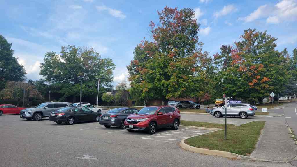A parking lot filled with various cars surrounded by trees in early autumn, showcasing colorful foliage against a partly cloudy sky.