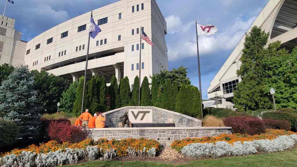 Virginia Tech campus entrance with the 'VT' logo, surrounded by landscaped flowers and greenery, and featuring several flags, including the American flag.