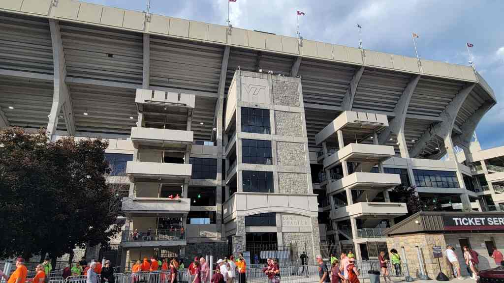 Exterior view of a large sports stadium with a modern design, featuring multiple levels, a stone facade, and visible stadium signs.