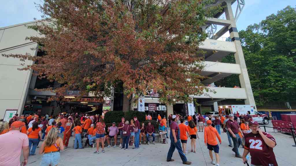 A crowd of fans wearing orange and maroon apparel gathered outside a stadium with a large tree in the foreground, showing autumn foliage.