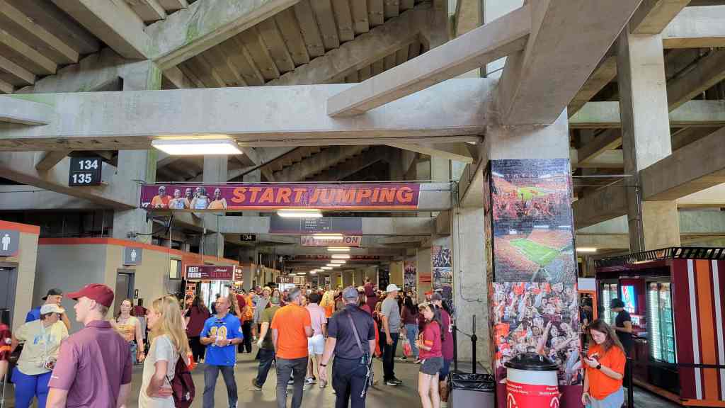 Crowd of people walking in a stadium corridor, with signs indicating sections and restrooms. A banner reads 'START JUMPING' above the walkway.