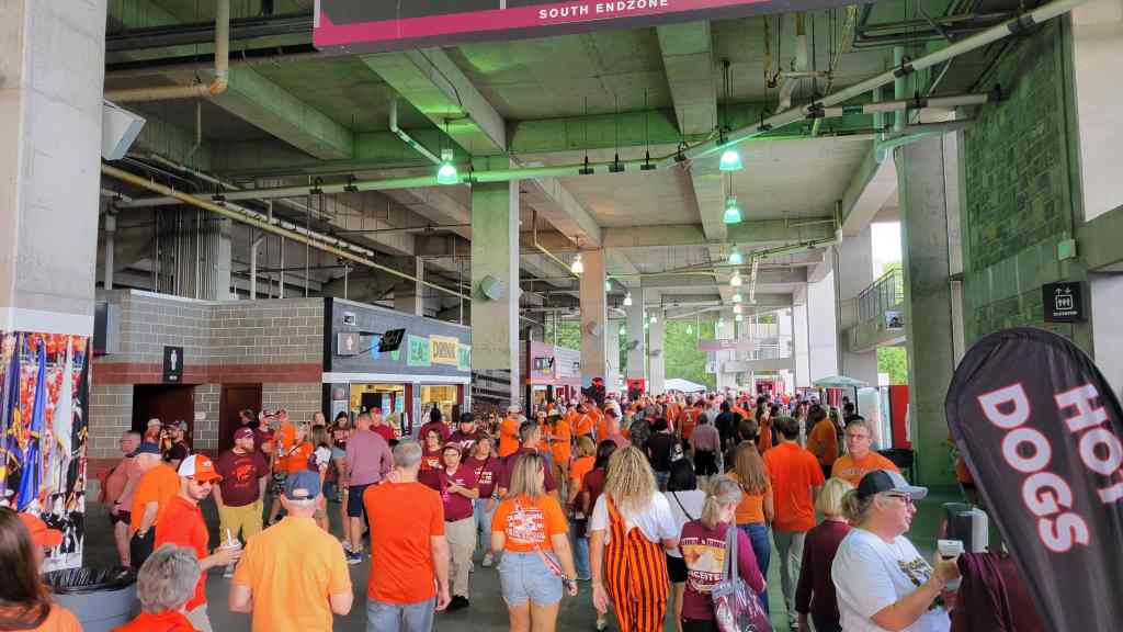 A bustling concourse filled with fans in orange and maroon clothing, enjoying refreshments and merchandise stalls in a sports venue.