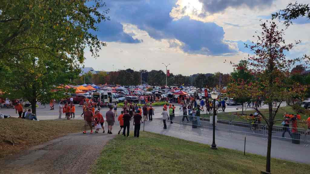 A busy outdoor event scene with many people walking in a parking area filled with tailgating setups, orange tents, and vehicles under a partly cloudy sky.
