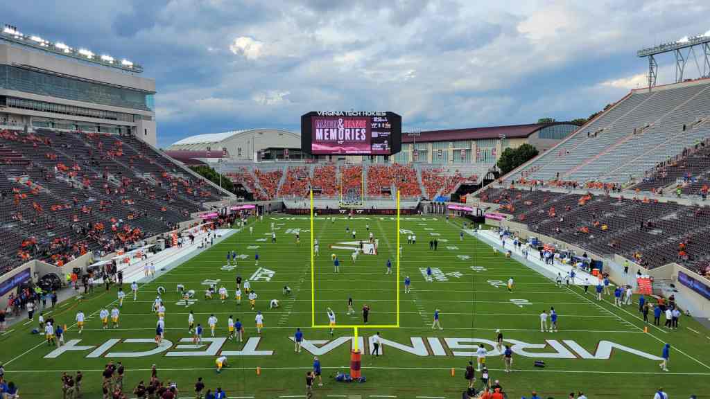 View of Virginia Tech football stadium during warm-ups, featuring players on the field and a large digital scoreboard displaying 'Memories'.