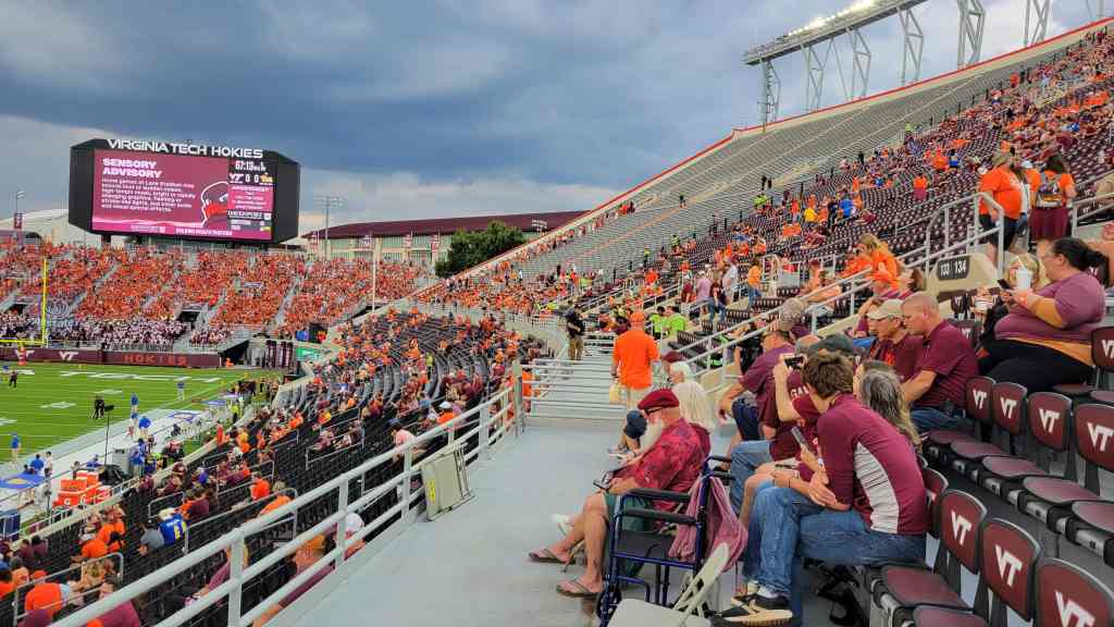 A partially filled stadium during a Virginia Tech football game, with fans wearing orange and maroon colors. The large scoreboard displays a sensory advisory message, and dark clouds loom in the sky.