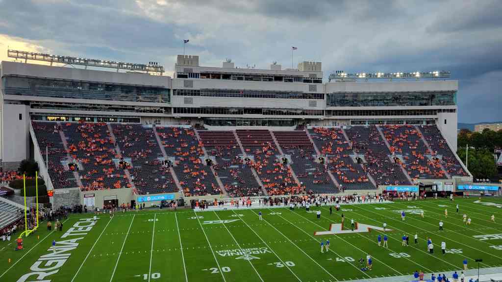 A view of a football stadium with empty and partially filled seating, a green field marked with yard lines, and a cloudy sky in the background.