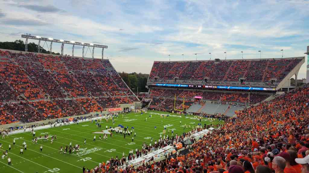 A crowded football stadium filled with fans wearing orange and maroon, with players warming up on the field under a cloudy sky.