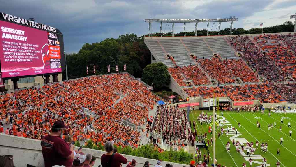 A large crowd of Virginia Tech fans wearing orange and maroon fills the stands at Lane Stadium during a football game. A big screen displays a sensory advisory, with players and the marching band visible on the field.