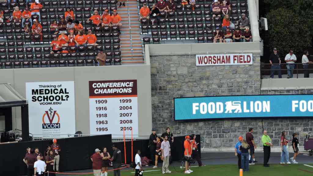 View of Worsham Field with spectators wearing orange and maroon in the stands, banners displaying conference championships, and advertisements for Food Lion.