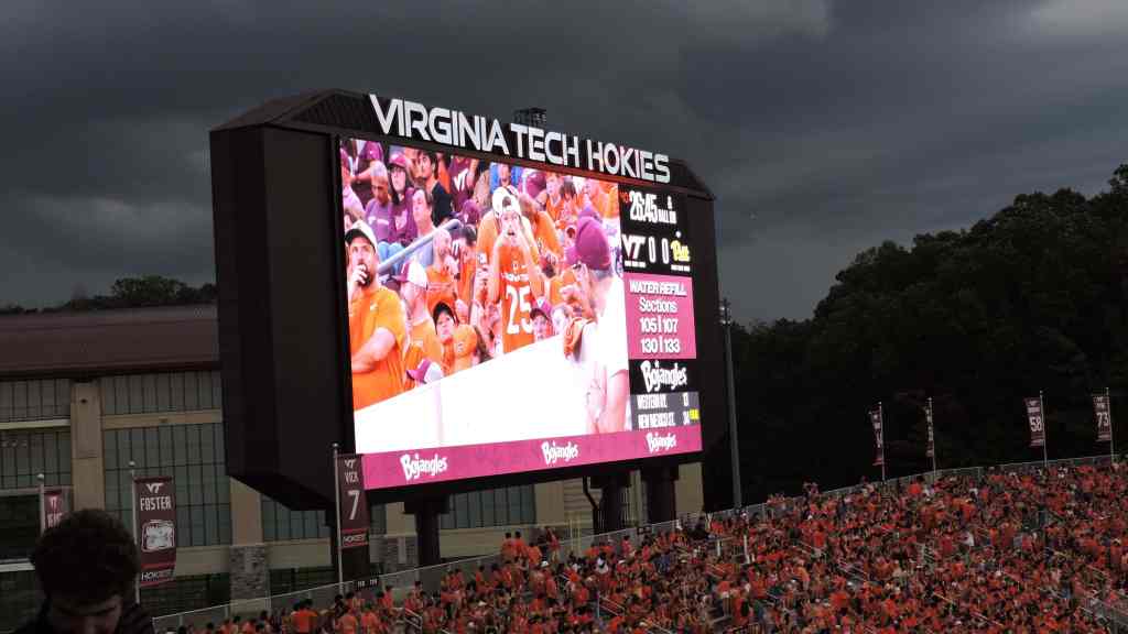 A large scoreboard displays the Virginia Tech Hokies logo and game information in a stadium filled with fans wearing orange apparel, with dark clouds in the background.