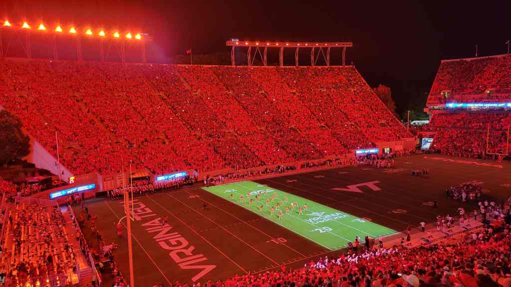 A crowded stadium at night, filled with fans wearing orange and maroon, with bright orange lights illuminating the scene and a football field in the foreground.