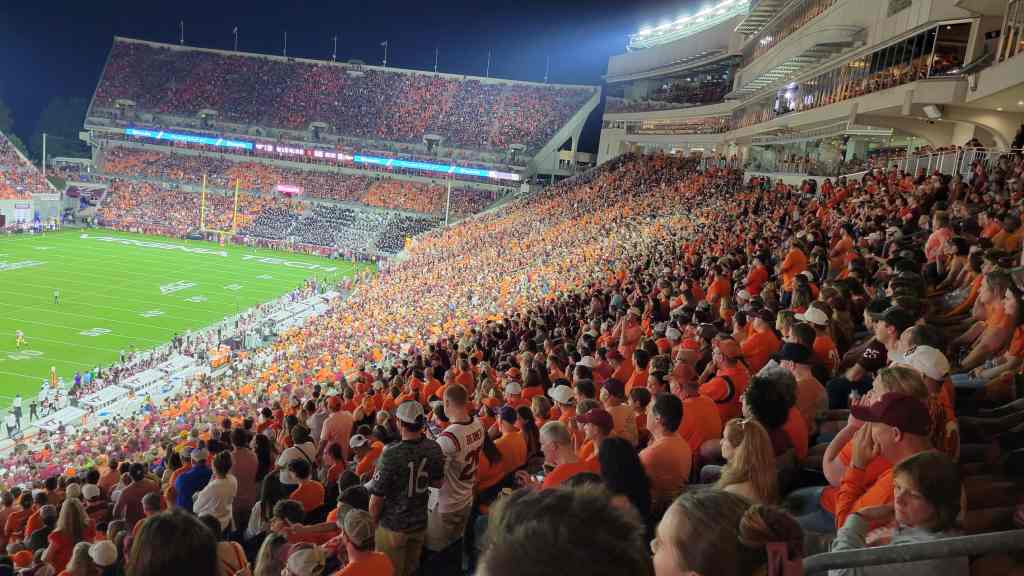 A crowded football stadium filled with fans wearing orange shirts, with the field visible in the background under stadium lights.