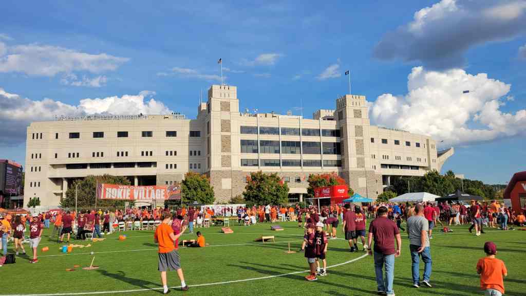 A lively scene at Hokie Village with crowds of fans wearing maroon and orange, enjoying festivities in front of a large stadium under a bright blue sky with clouds.